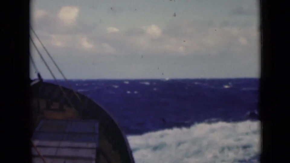 1947 8mm view from a boat deck, with the bow and rigging pointed toward rough blue water and low clouds.