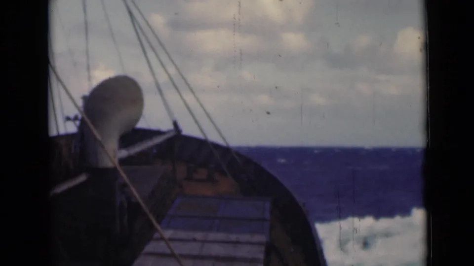 The boat’s wooden forward deck and taut rigging appear against a cloudy horizon and dark blue, wind-ruffled sea.