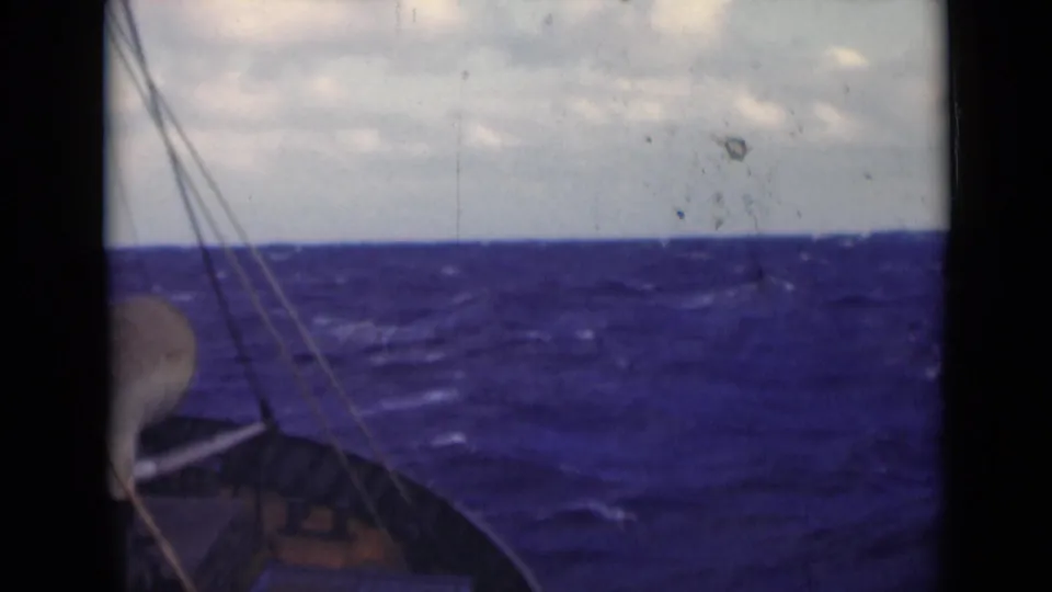 A handheld 8mm frame shows rigging, part of the deck, rough sea surface, and a cloudy sky during a small-vessel passage.