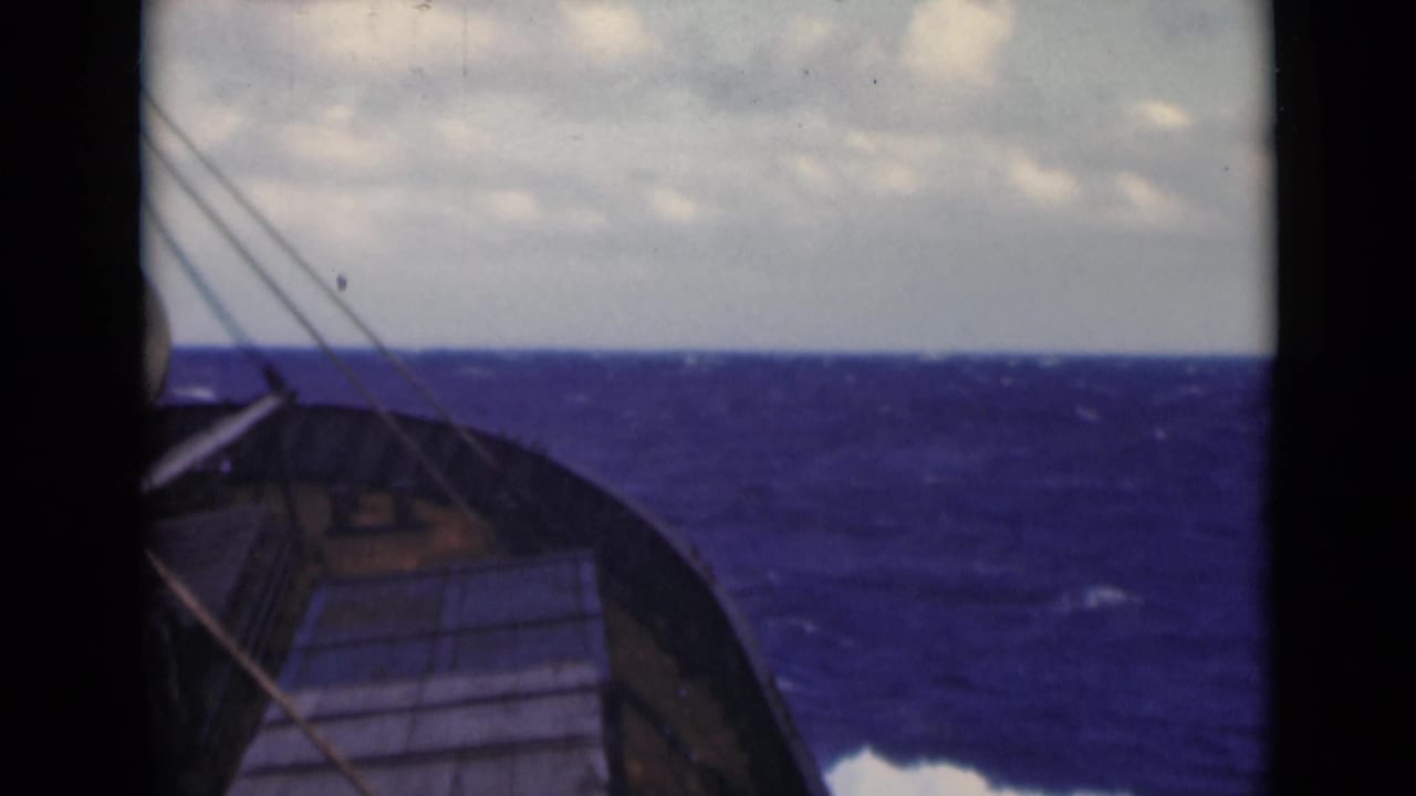 Deck-level 8mm view of a ship bow and rigging facing rough blue water, with an overcast horizon visible beyond the rail.