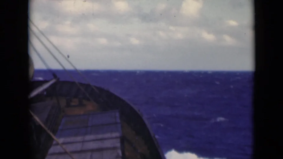 Deck-level 8mm view of a ship bow and rigging facing rough blue water, with an overcast horizon visible beyond the rail.