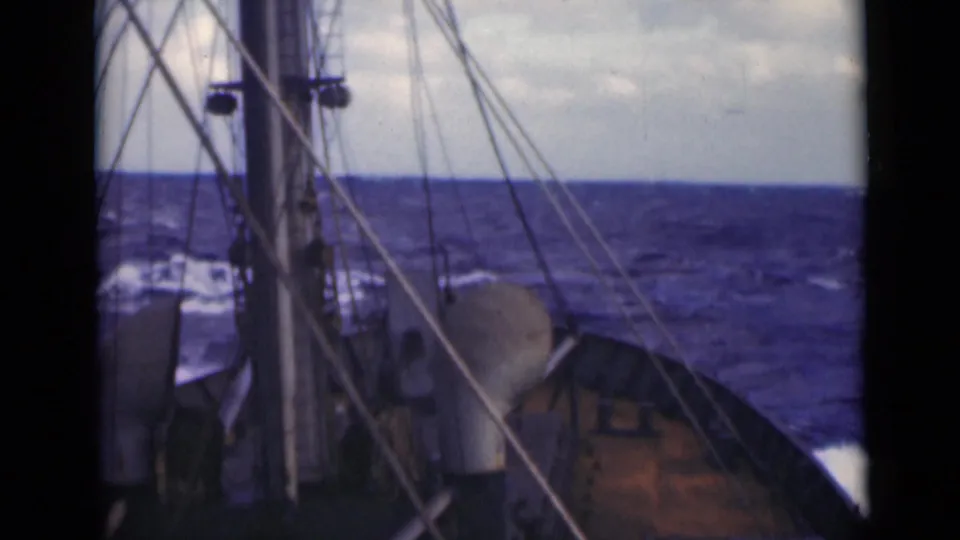 A mast and rigging dominate the deck view as the vessel moves across choppy water, with a cloudy horizon in the background.