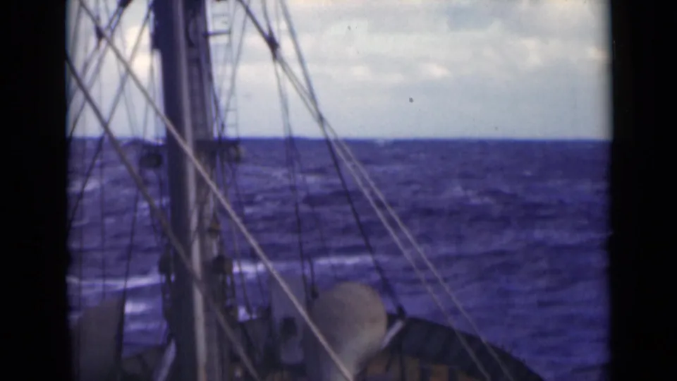 The frame centers on the ship’s mast and rigging, with dark blue rough water and a cloudy sky beyond the deck.