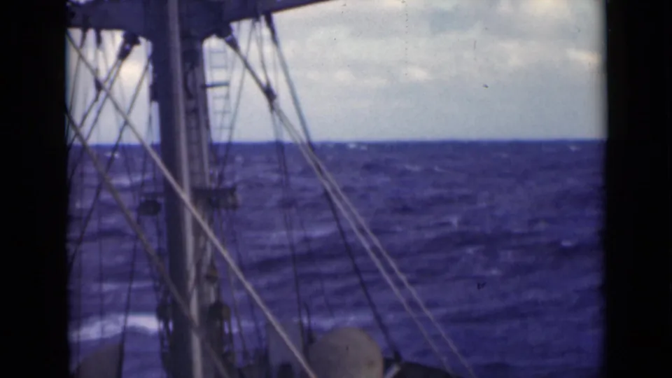 A deck-level view shows mast hardware and rigging in front of rough open water, with the ship’s movement implied by the tilted framing.