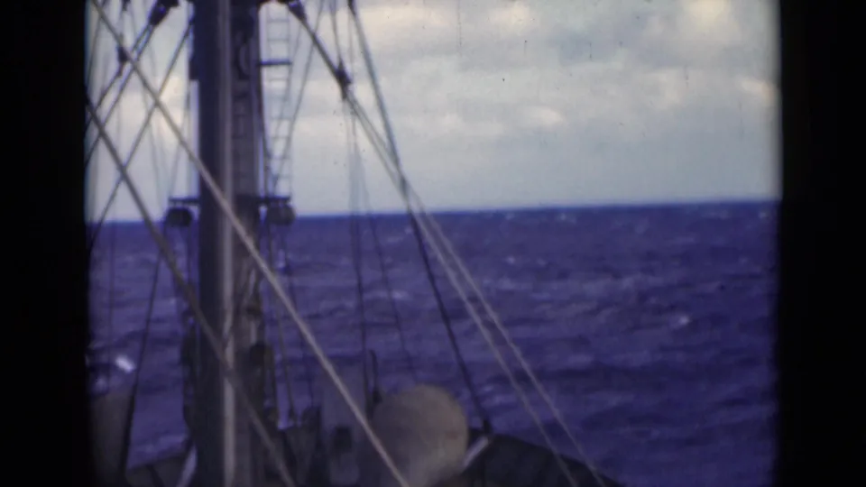 1947 8mm boat-deck view with rigging, mast hardware, and turbulent blue water below a clouded sky.