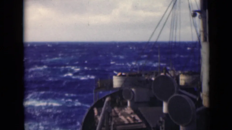 A ship's forward deck and circular fittings sit beside rough blue sea, with rigging lines crossing the overcast horizon.