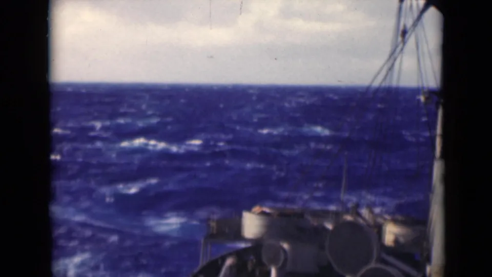 The camera looks past the ship's edge toward dark blue waves and whitecaps, with rigging and deck hardware partly visible.