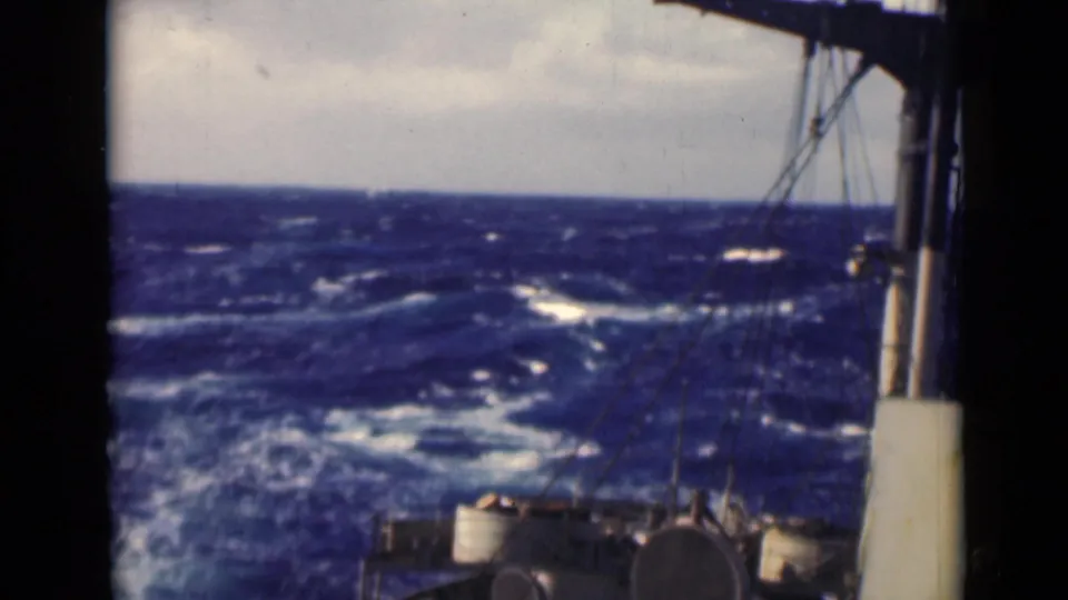 Deck-facing view from a vessel in rough water, with rigging, cylindrical fittings, and an overcast horizon.