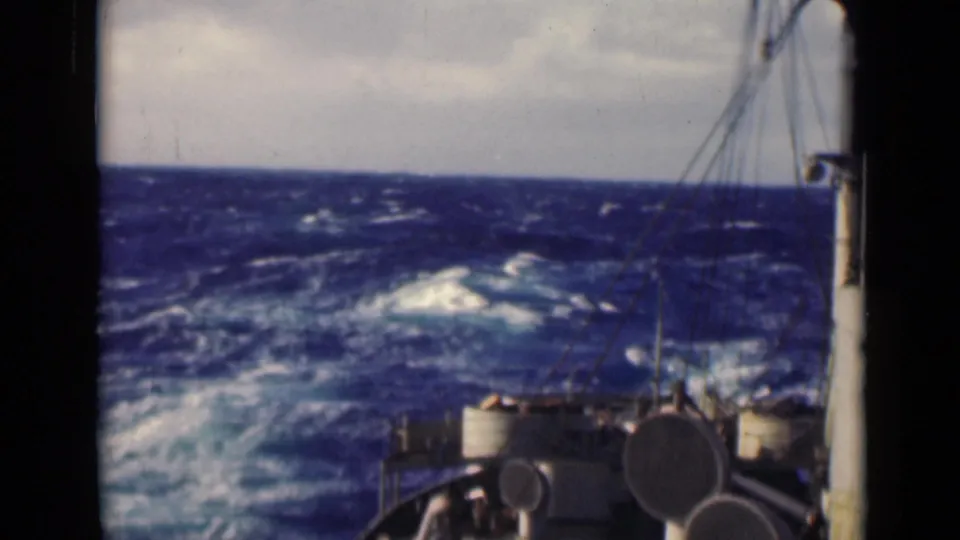 Vintage 8mm frame showing a vessel deck in the foreground as rough blue water rises toward a cloudy horizon.