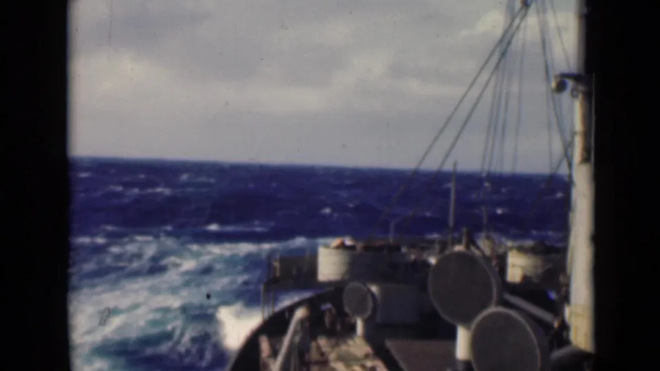 Rough sea and foaming water pass alongside visible deck equipment and rigging on a military-associated vessel.