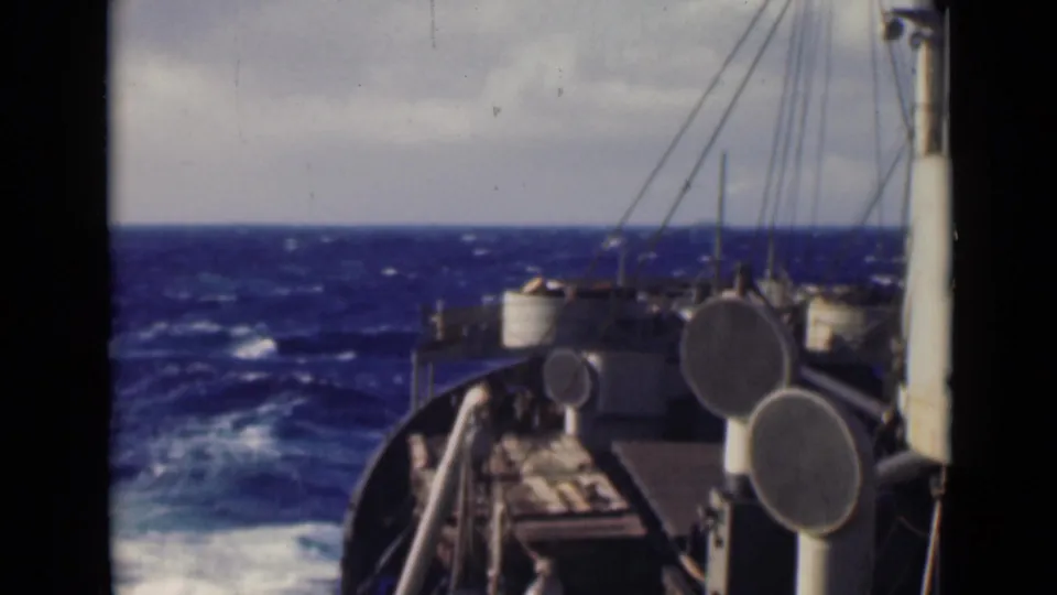A slightly lower forward view shows the ship bow, deck hardware, circular ventilators, rigging lines, and white spray beside deep blue waves.