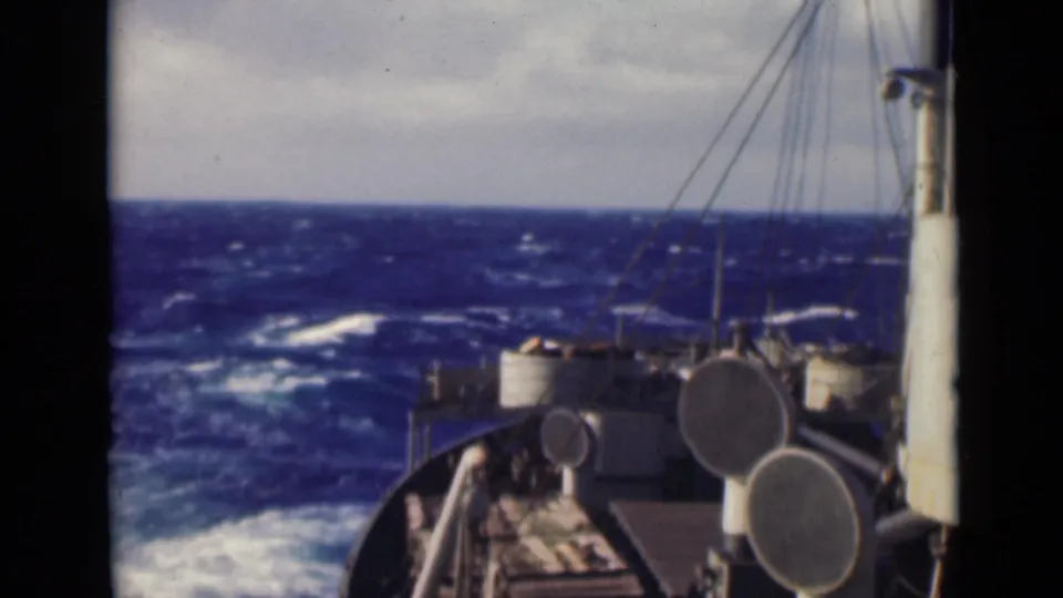The ship's bow and deck fittings remain in view as rough open water and white wake fill the background under a gray sky.