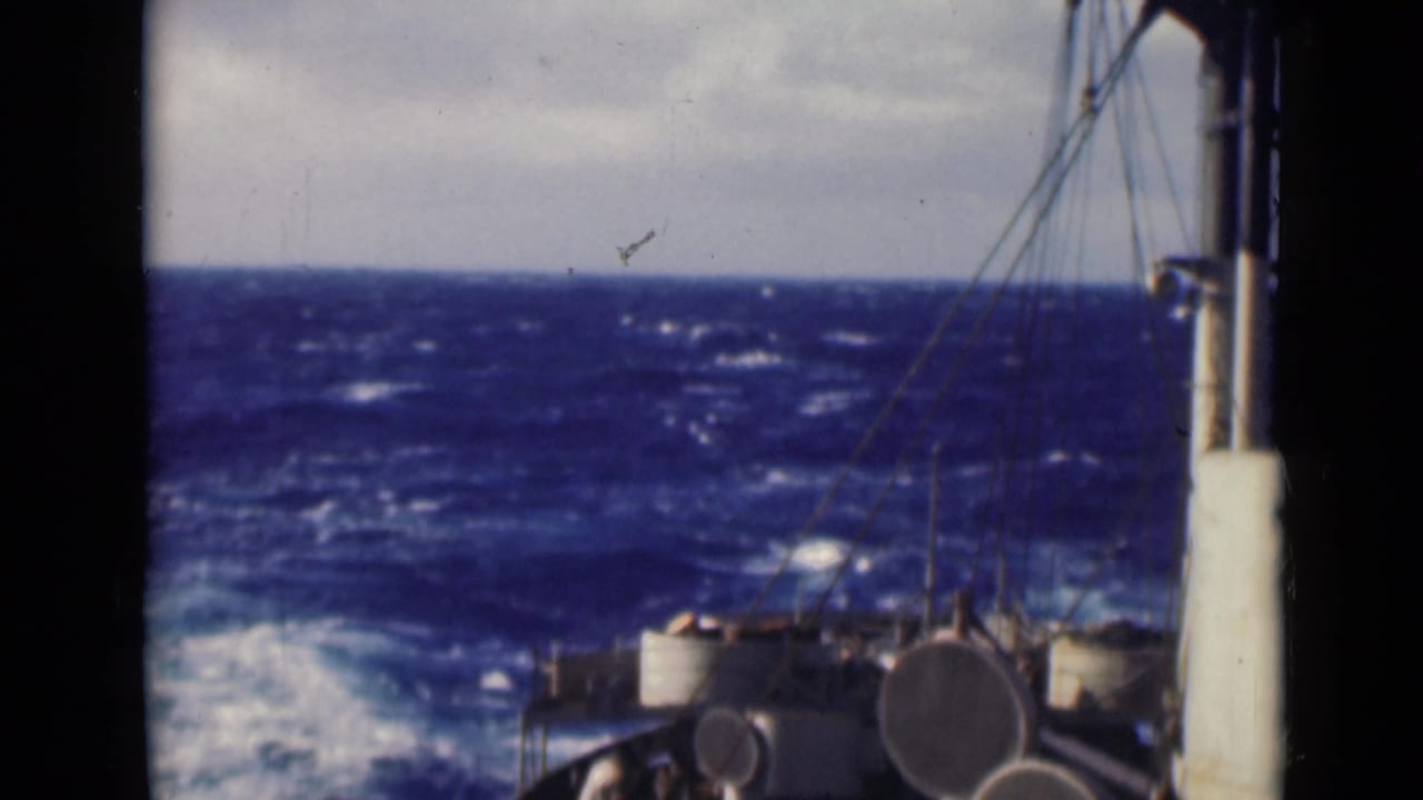 Shipboard view across the bow toward rough blue waves, with railings, rigging, and deck equipment partly obscured by film-frame edges.