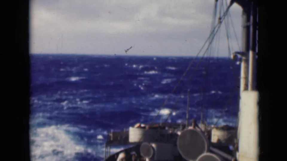 Shipboard view across the bow toward rough blue waves, with railings, rigging, and deck equipment partly obscured by film-frame edges.