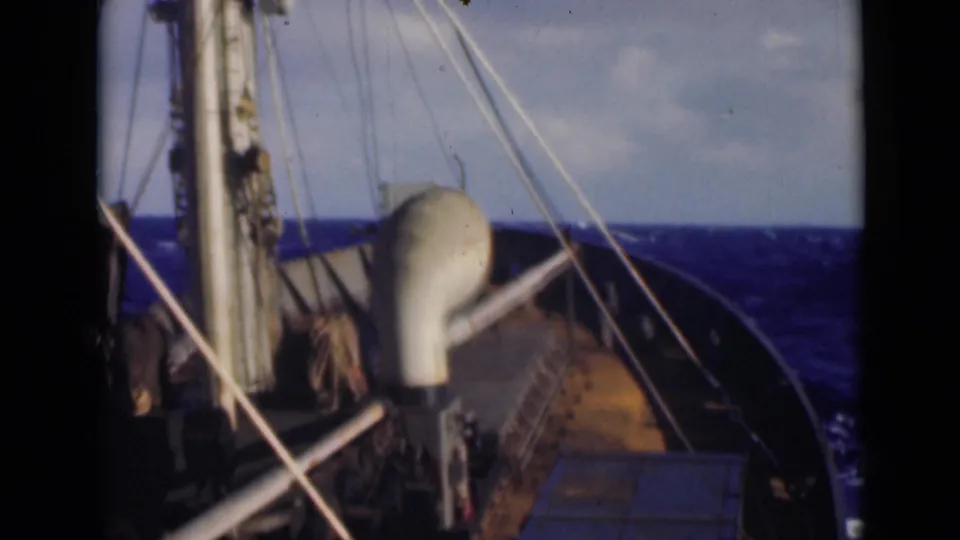 View along the ship's forward deck with mast, rigging, railings, and a vent-like fitting as rough blue water surrounds the bow.