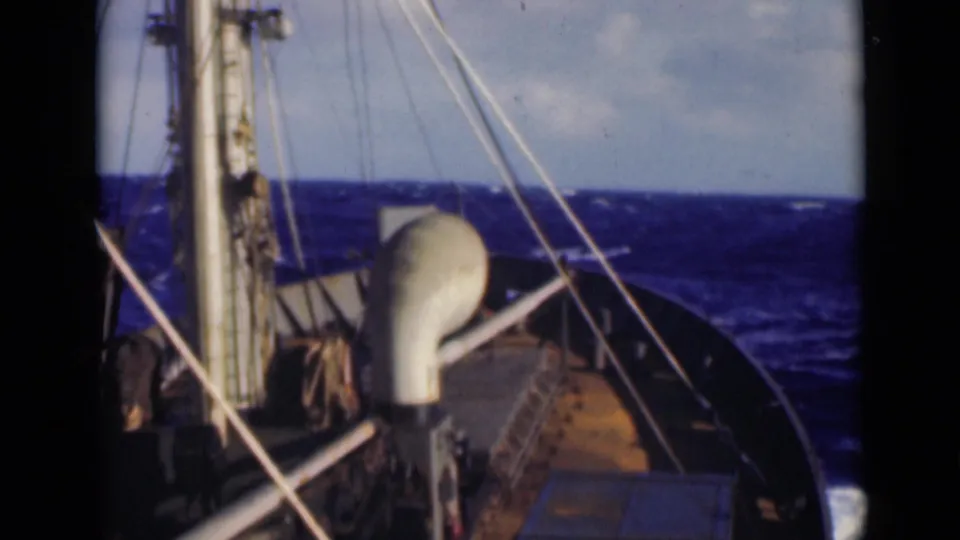 1947 8mm frame from ship deck with mast, rigging, white deck structure, and rough blue sea ahead.