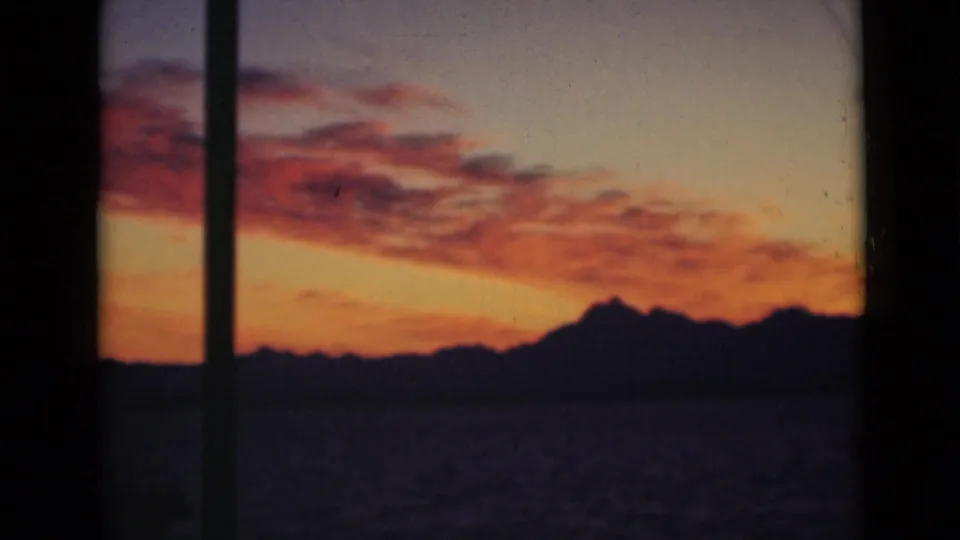 Color 8mm frame showing a sunset cloud band over water and a dark, mountain-like horizon, partially blocked by window framing.