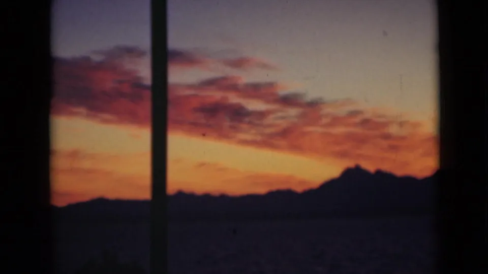 Home-movie frame of a warm sunset sky, dark water, and a silhouetted horizon behind vertical window framing.