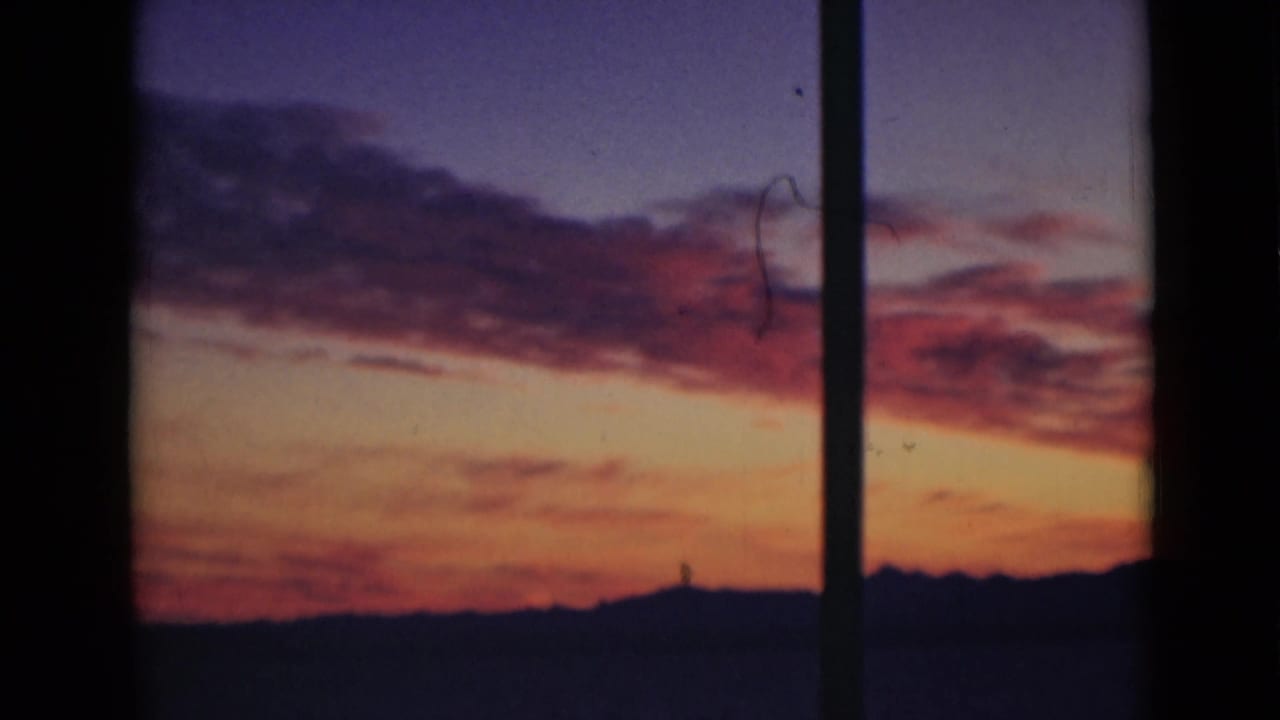 Archival 8mm view of red-purple clouds over an orange sunset horizon, framed by dark vertical bars in the foreground.