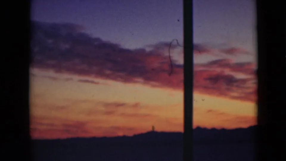 Archival 8mm view of red-purple clouds over an orange sunset horizon, framed by dark vertical bars in the foreground.