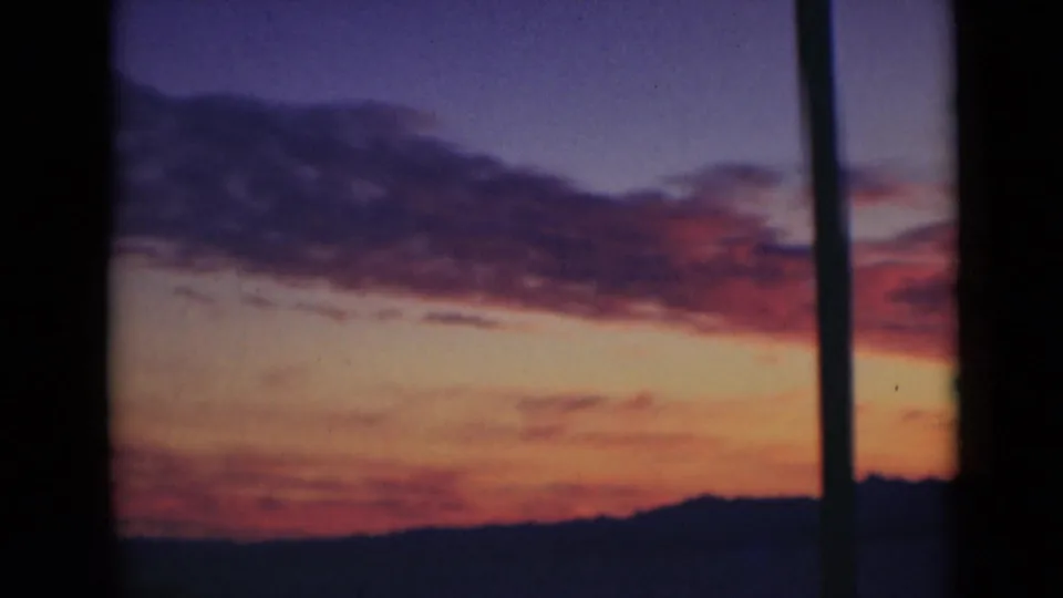 1947 home-movie sunset frame with a broad red cloud band, orange lower sky, dark horizon, and a vertical pole near the right edge.