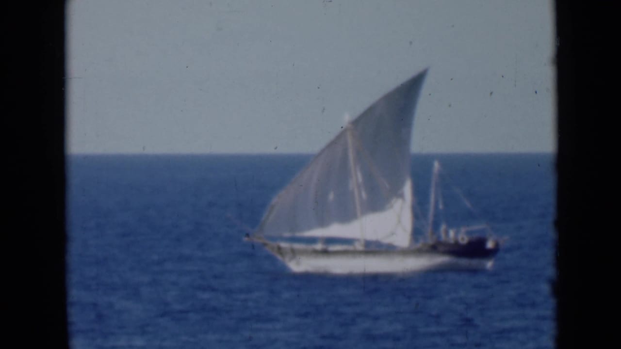White-sailed dhow-like boat crossing blue water under a pale sky.