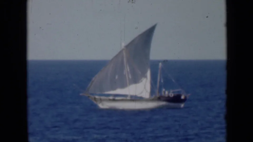 Soft-focus 8mm frame of a dhow-like vessel with a raised white sail, seen broadside against blue water and a clear horizon.