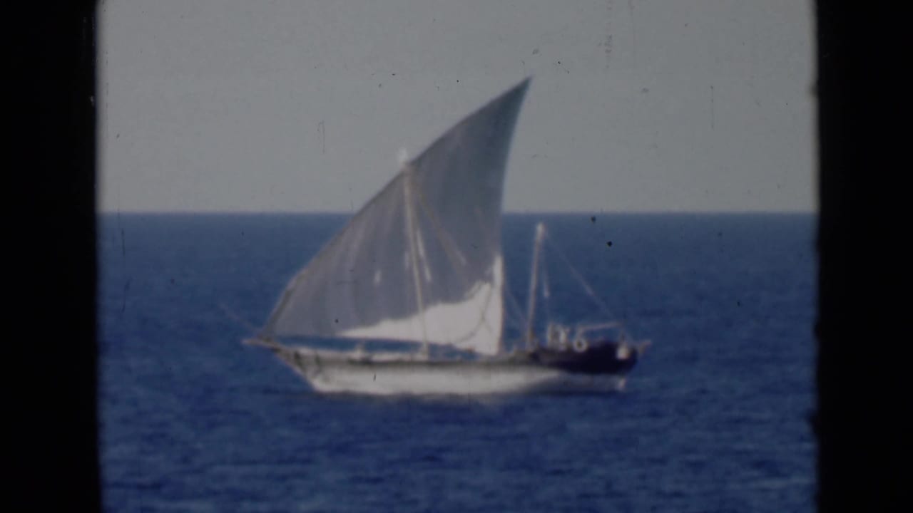 A traditional dhow appears at sea with a large white lateen sail, seen in soft 8mm footage against a pale sky and blue water.
