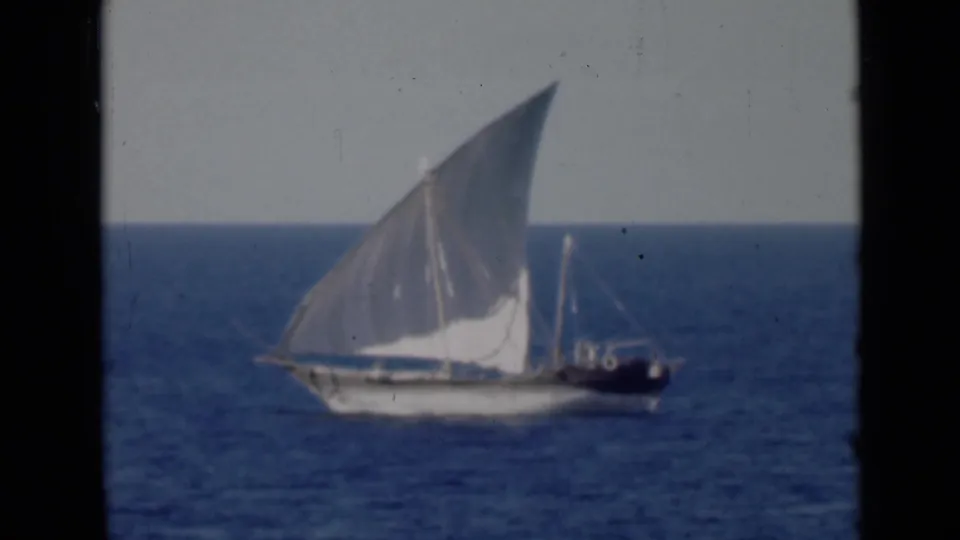 A traditional dhow appears at sea with a large white lateen sail, seen in soft 8mm footage against a pale sky and blue water.