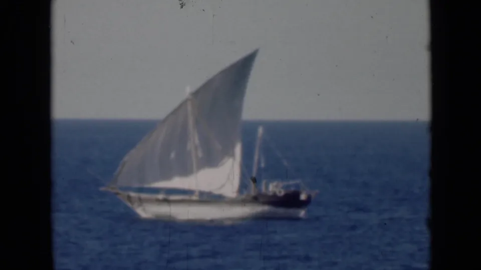 The dhow remains in profile on the Persian Gulf, its white sail catching light as the boat moves across calm blue water.