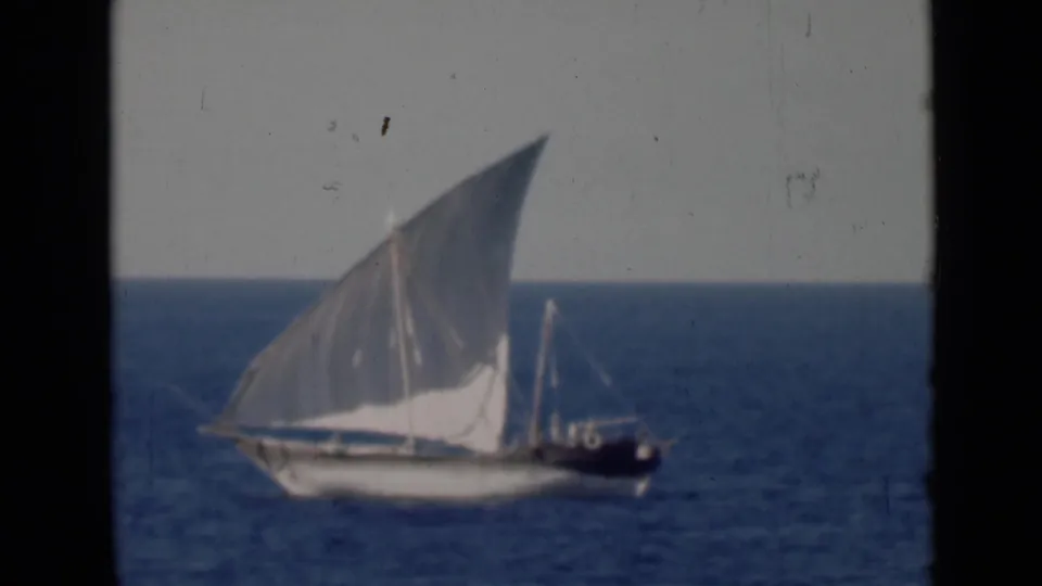 A soft 8mm frame shows the dhow lower against the blue sea, with a broad white lateen sail and a pale sky above the horizon.