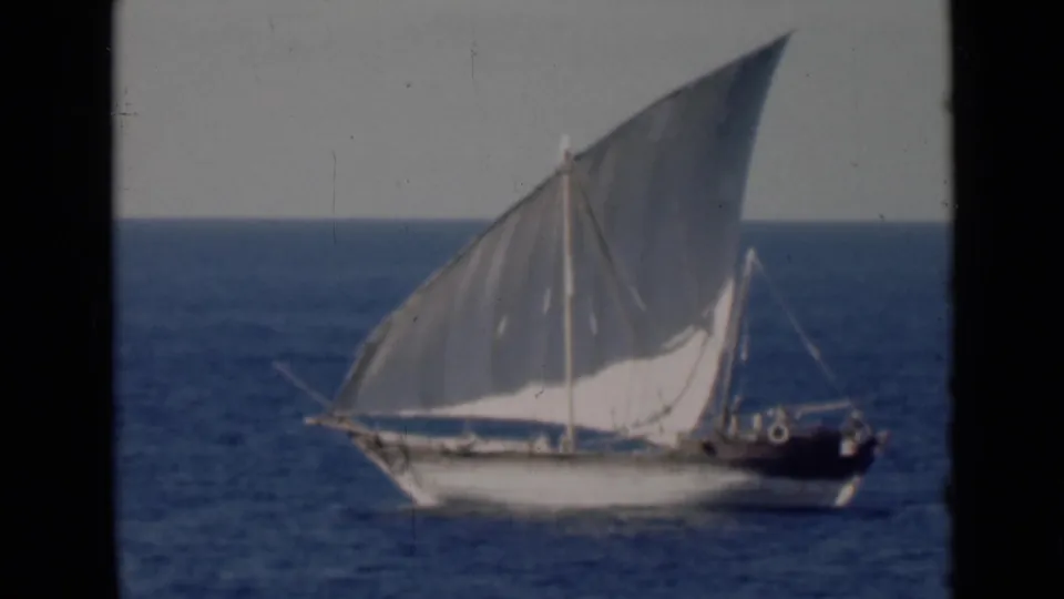 The final frame shows the dhow slightly closer, with its large white sail, small hull, and dark stern set against blue water.