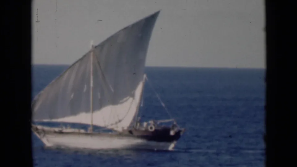 A cropped view shows the dhow passing through frame, with rigging, dark stern area, and possible small figures aboard.