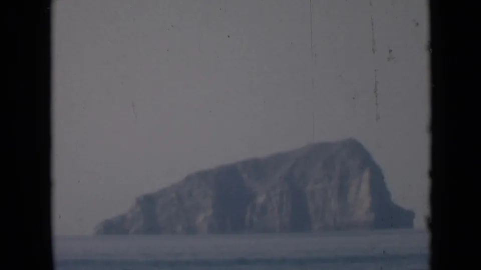 A hazy rocky island appears on the horizon beyond calm water, closing the short maritime 8mm sequence.