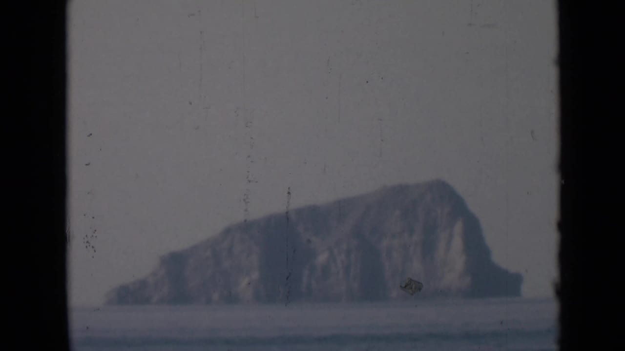 Distant rocky island across calm water, framed by dark vertical film or window edges.