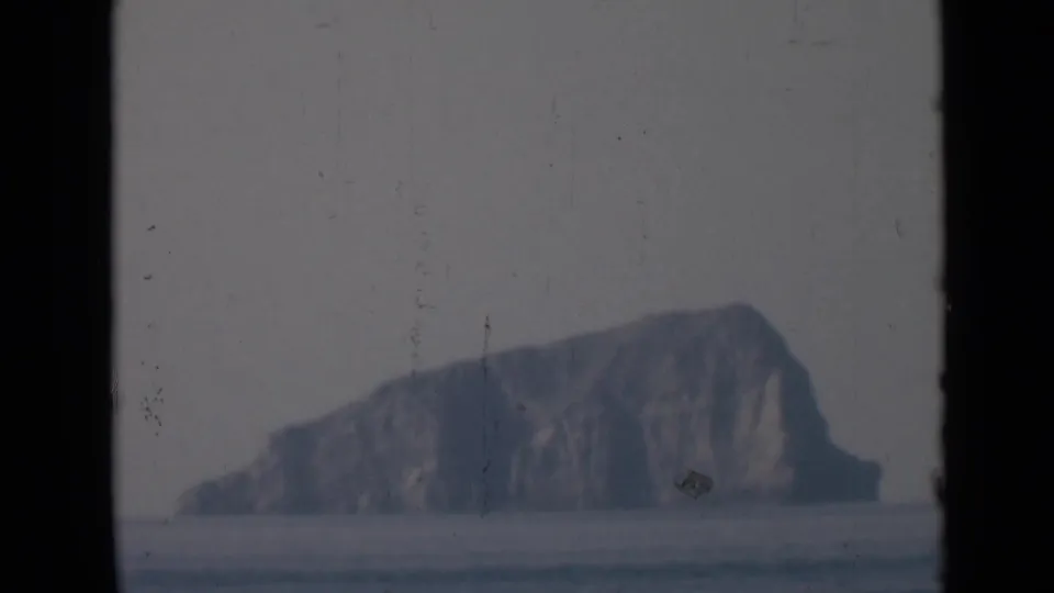 A hazy offshore rocky island sits beyond calm sea, with dark vertical edges suggesting filming through a window or porthole.