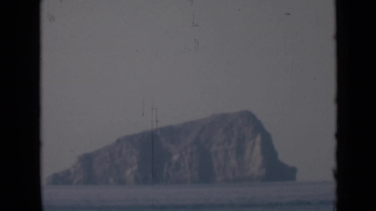 The rocky island remains centered beyond flat water, softened by haze and framed by dark vertical borders from the shooting position.