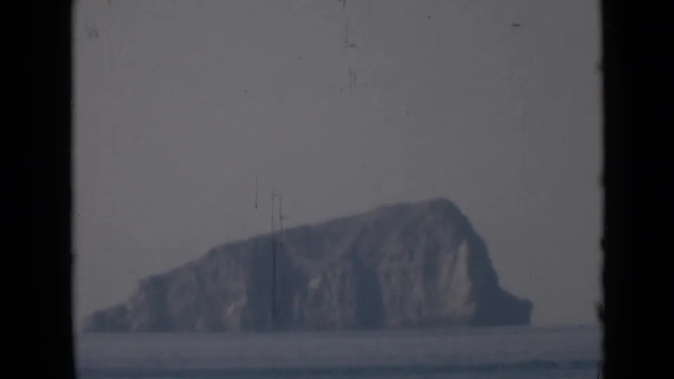The rocky island remains centered beyond flat water, softened by haze and framed by dark vertical borders from the shooting position.