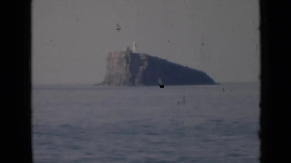 A lower rocky island appears across calm sea, with a small light-colored tower or lighthouse-like marker visible near the top.