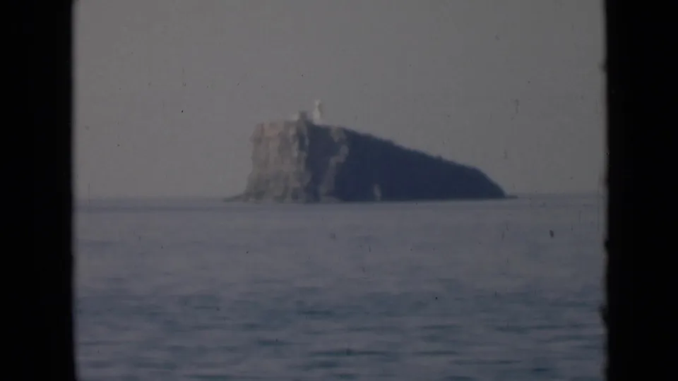 The final frame shows the rocky island and a small pale lighthouse-like structure against a hazy sky above the quiet sea.