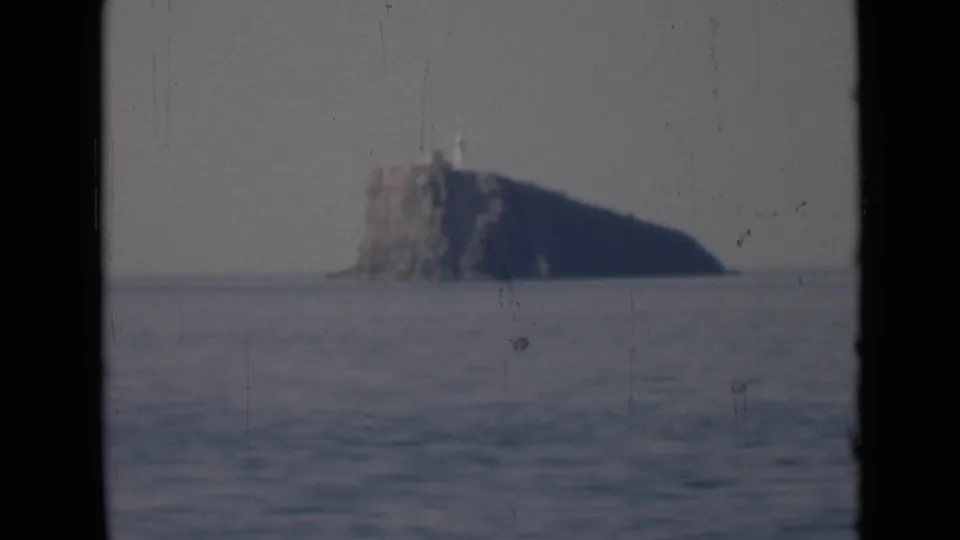 1947 8mm frame showing a distant rocky island or sea stack with a lighthouse-like structure above calm water in the Persian Gulf region.