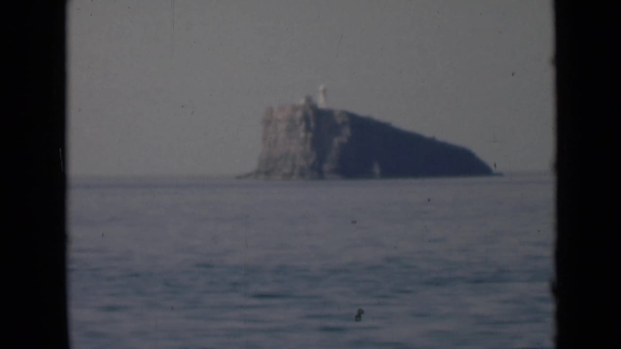 A distant rocky island with a lighthouse-like structure is seen through dark film borders, with open water in the foreground.