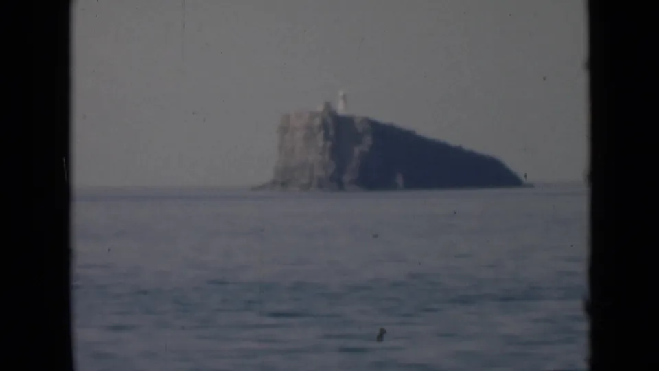 A distant rocky island with a lighthouse-like structure is seen through dark film borders, with open water in the foreground.