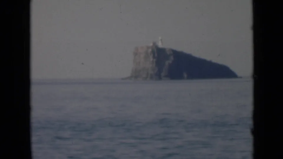 Vintage 8mm view of a low rocky island or sea stack with a small light structure, filmed from across blue-gray water.