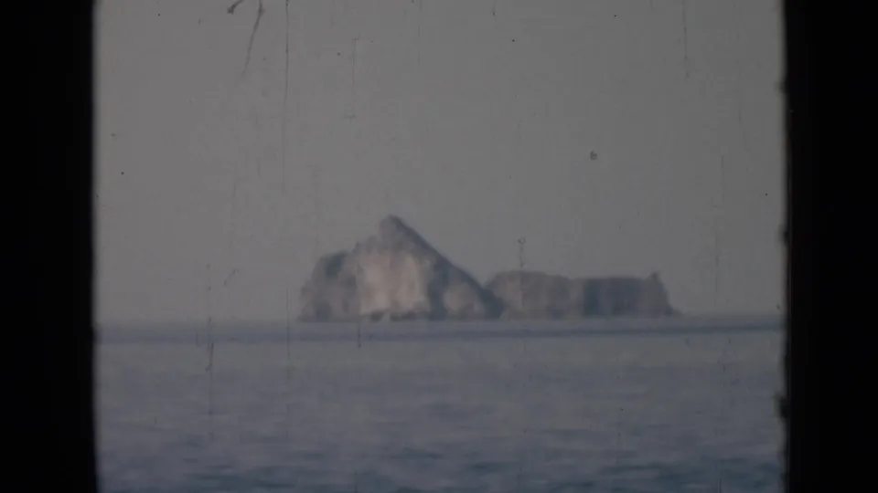 Soft 8mm view of a distant rocky island across calm sea under a hazy sky, with dark frame borders visible.
