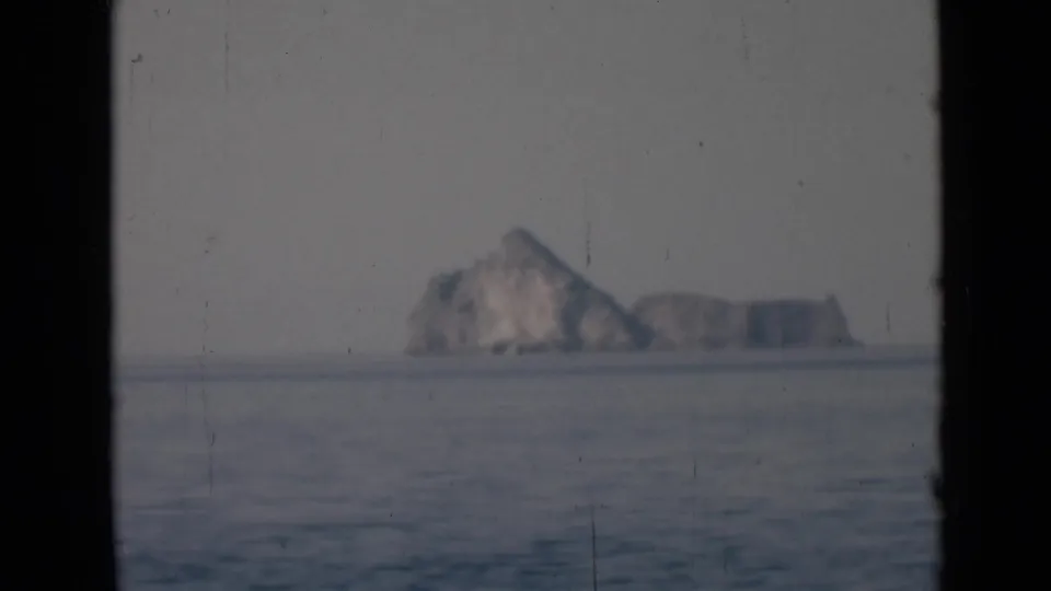 Offshore home-movie view of a rocky island rising from the horizon, with calm water and visible 8mm grain.