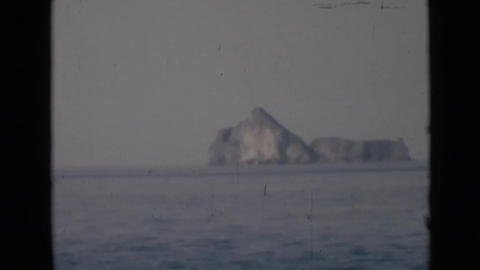 A far rocky island and lower ridge sit beyond calm ocean water, framed by dark edges and a hazy sky.