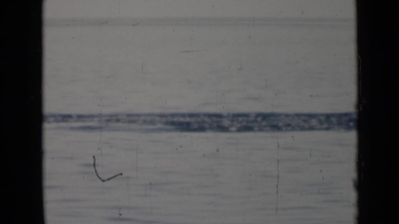 Archival 8mm frame of calm open water and a distant low shoreline, partly divided by a dark window mullion.