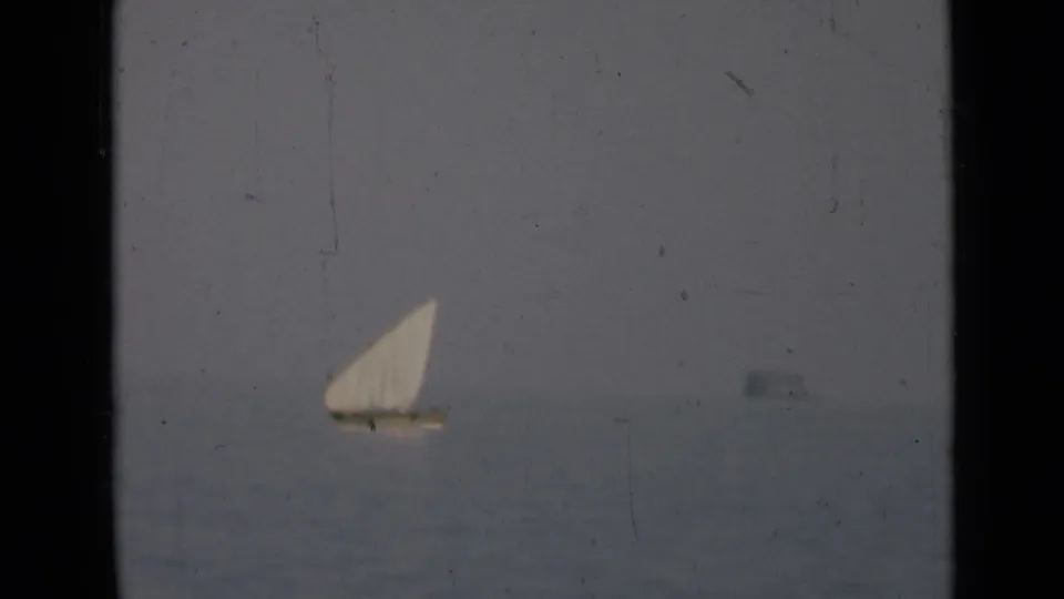 A distant traditional sailboat appears on calm water under a hazy sky in archival 8mm footage associated with the Persian Gulf.