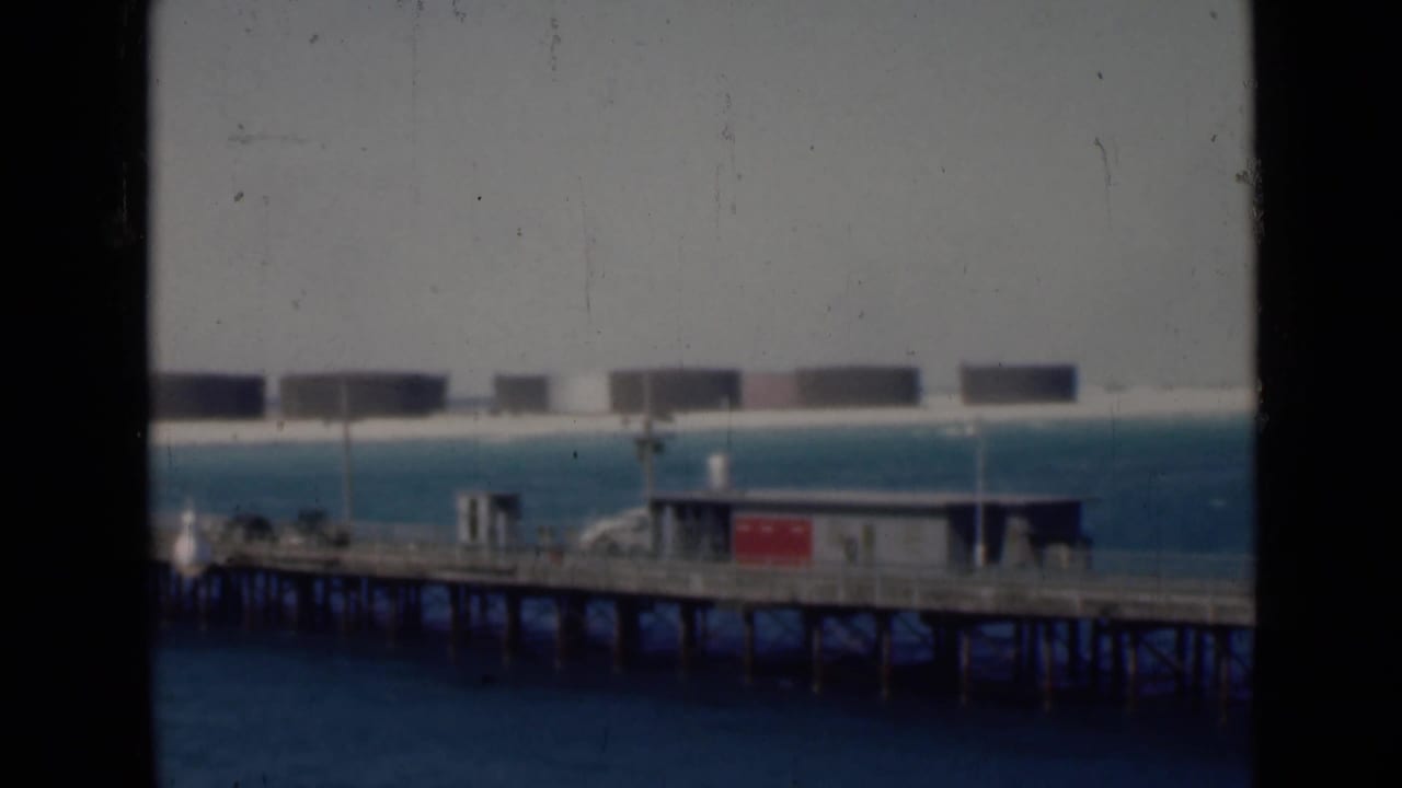 Wooden pier over blue water with small buildings and distant storage tanks.